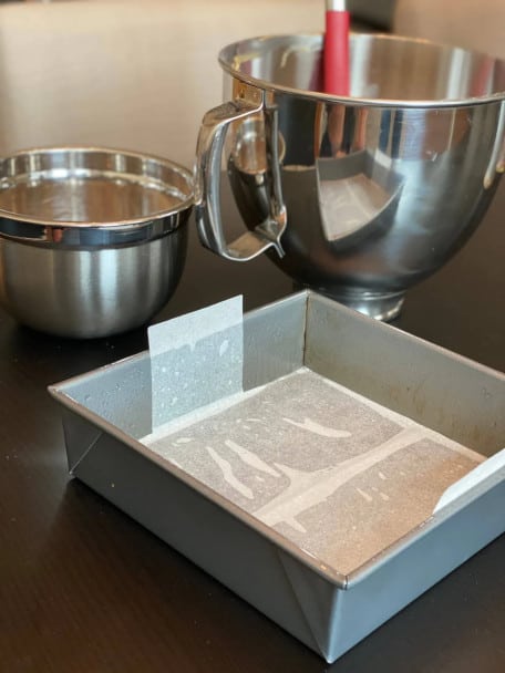 A square baking pane lined with tabbed parchment, mixing bowls in the background