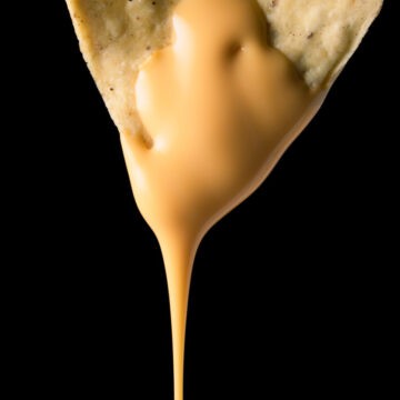Closeup photo of a nacho with liquid cheese dripping from it on a black background